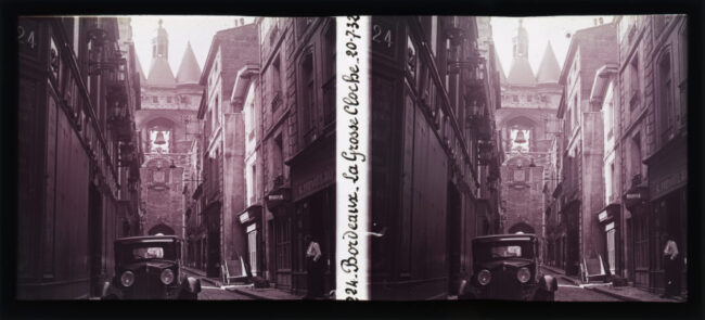 Vue de la Grosse Cloche depuis la rue Saint-James. Une voiture ancienne est garée au premier plan, une charrette est visible au second plan. Au fond, au centre, se déploie l'édifice de la Grosse Cloche.