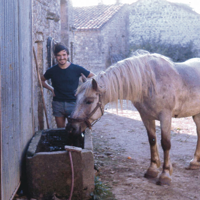 Au premier plan, une jument boit dans un bac en pierre au milieu d'une ferme. Au second, on peut voir un jeune homme souriant, regardant la scène.