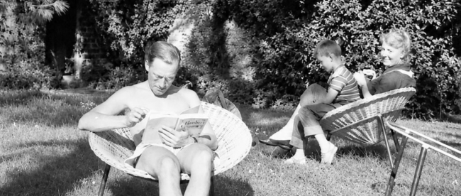 Photo en noir et blanc. On peut voir Michel Foucault au premier plan, assis, dans le jardin, en train de lire, et derrière sa mère également assise avec sur ses genoux le neveu de Michel Foucault